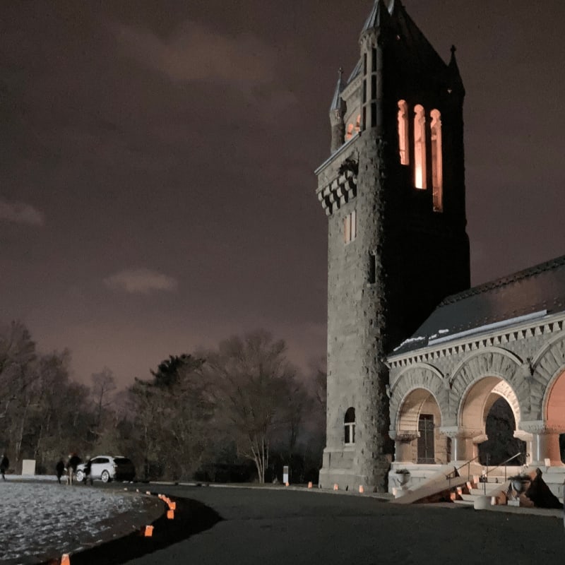 A tall stone tower with glowing windows stands beside a building with arched entryways, overlooking a dark paved area and a snowy landscape under a night sky.