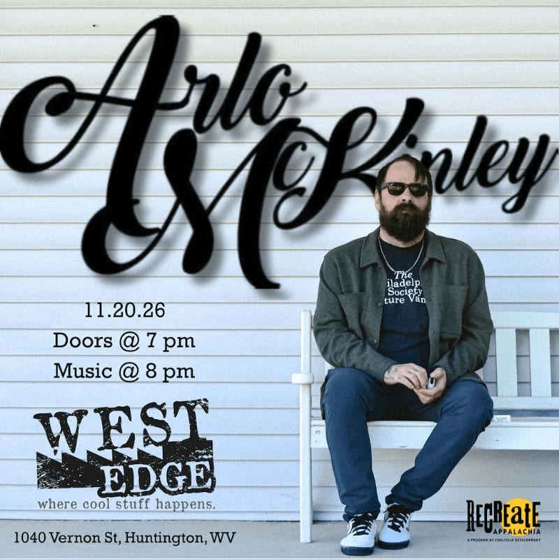 A bearded man in sunglasses and a grey jacket sits on a white bench in the foreground, set against a white siding wall with large black cursive text and event details in the background.