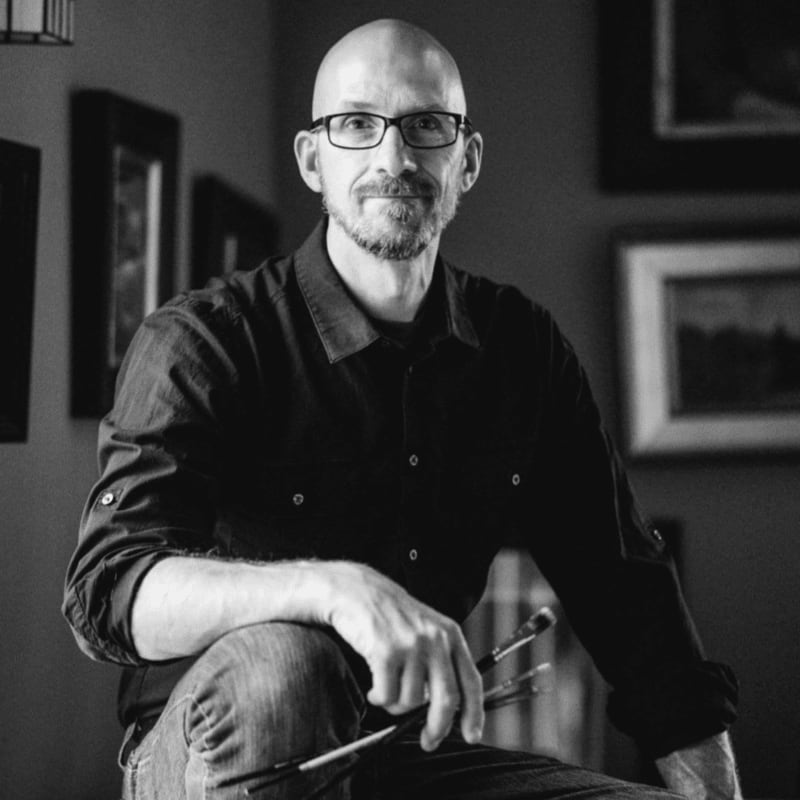 A bald man with glasses and a goatee wearing a black shirt holds paintbrushes in the foreground, set against a background of framed artwork on a wall.