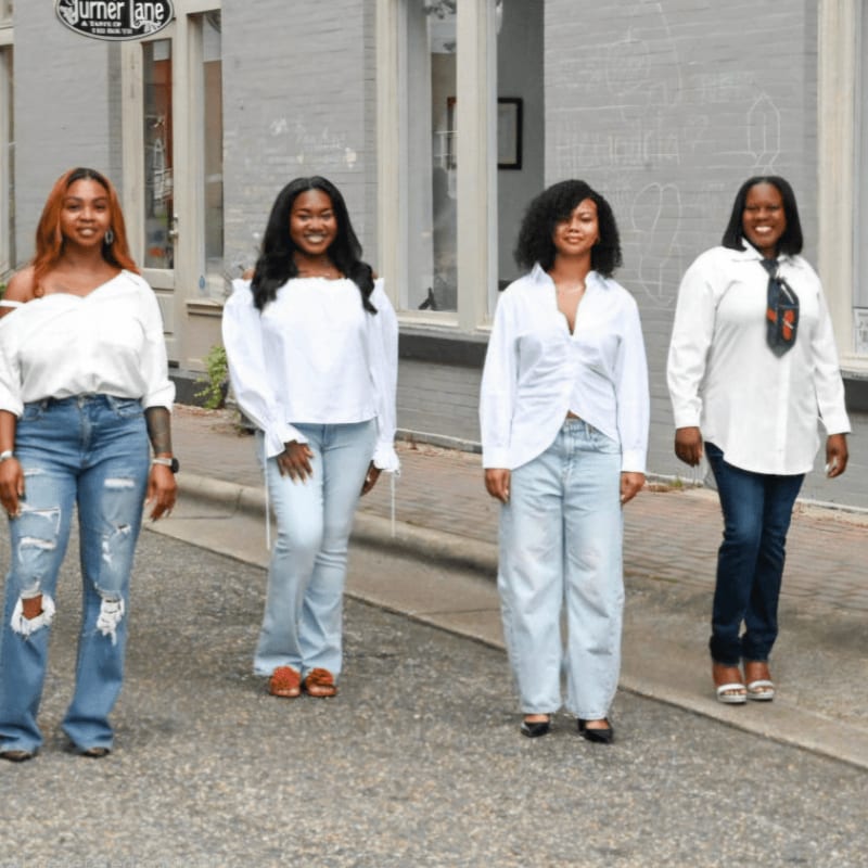 Four women wearing white tops and blue jeans stand on a paved street in front of a light-colored building.
