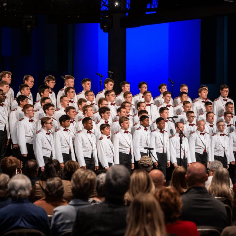 A choir of young men in white uniforms performs on a stage against a vibrant blue background, viewed from behind an audience.