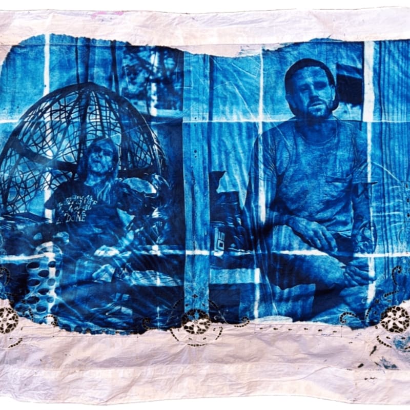 Two men are depicted in a blue cyanotype print, with one sitting inside a spherical cage in the foreground and the other sitting beside him against a textured background.