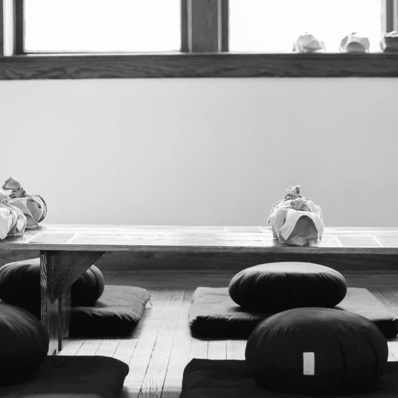 Round meditation cushions sit on floor mats in the foreground, while a long wooden bench with fabric bundles rests against a plain wall and window in the background.