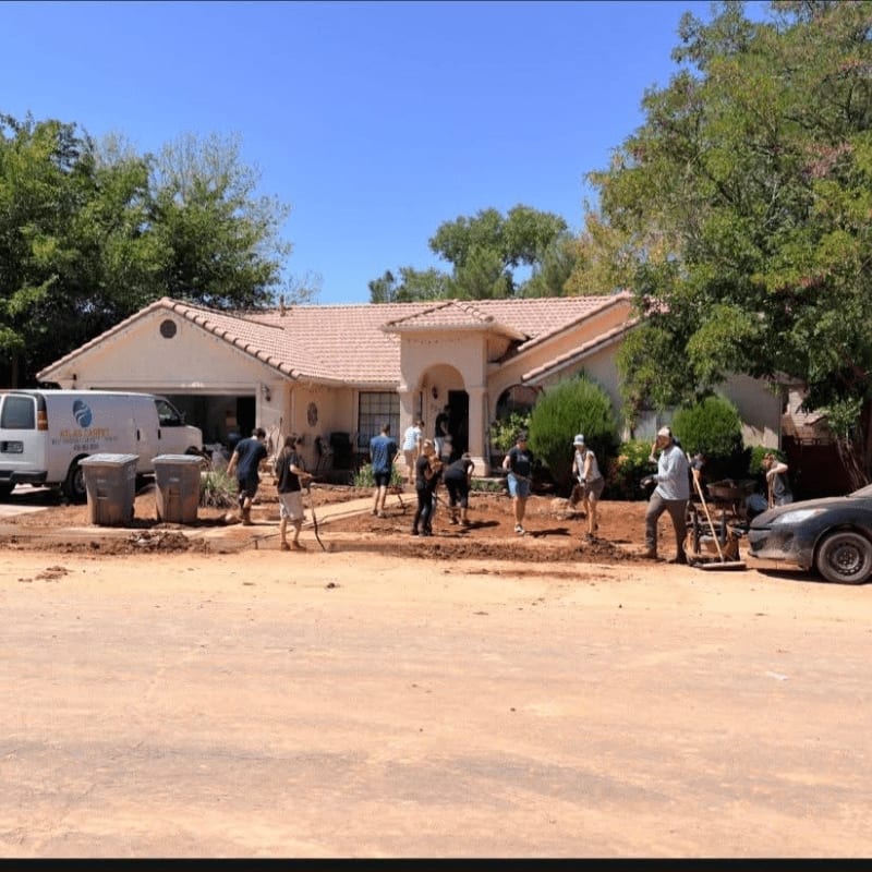 A group of people work on a dirt patch in the foreground in front of a single-story house and a white van under a clear blue sky.