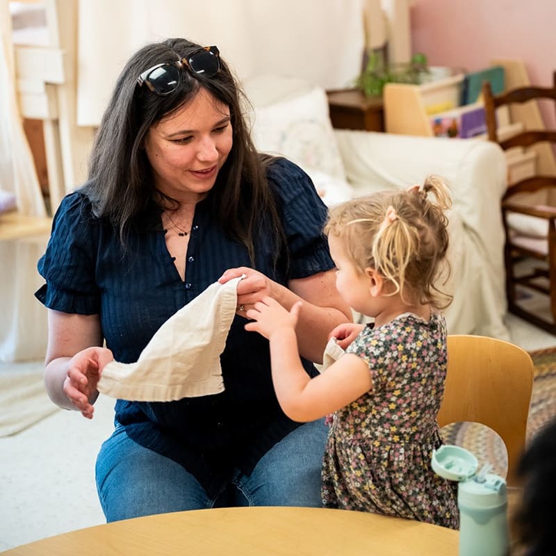 A woman in a dark blue shirt holds a piece of white fabric for a young girl in a floral dress, set within a cozy living room.