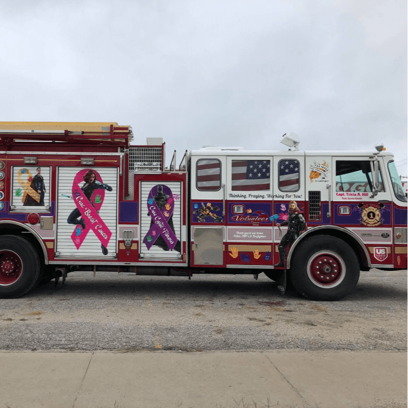 A red and white fire truck decorated with breast cancer awareness ribbons sits on a gravel lot under a cloudy sky.