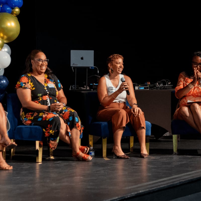 Three women sit in blue chairs on a stage during a panel discussion, with a dark background featuring a laptop and balloons.