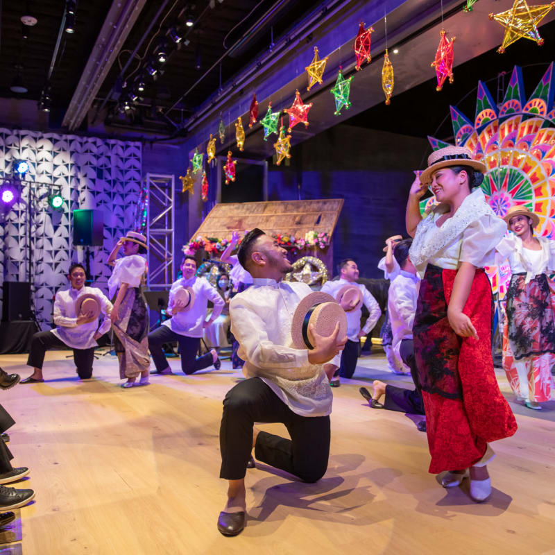 Performers in traditional Filipino attire dance on a wooden stage beneath colorful hanging stars and a vibrant decorative backdrop.