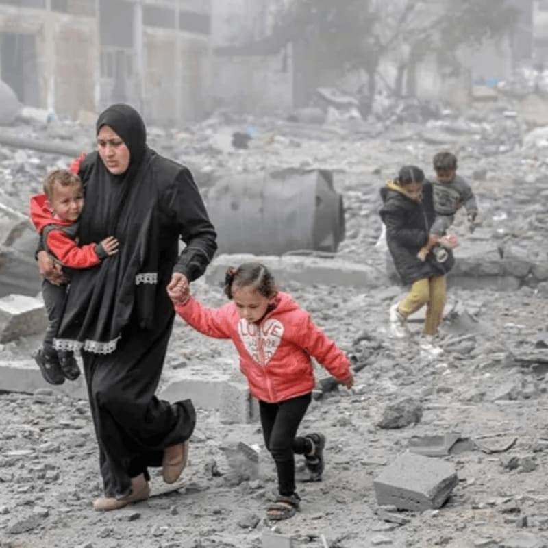 A woman in a black abaya carries a small child and holds the hand of a young girl in a red jacket amidst a landscape of grey rubble and ruins.