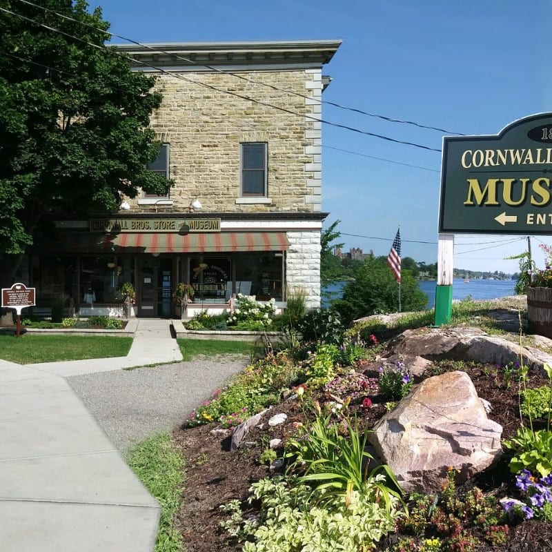 A flowerbed and a museum sign sit in the foreground before a stone building and a distant waterfront under a clear blue sky.