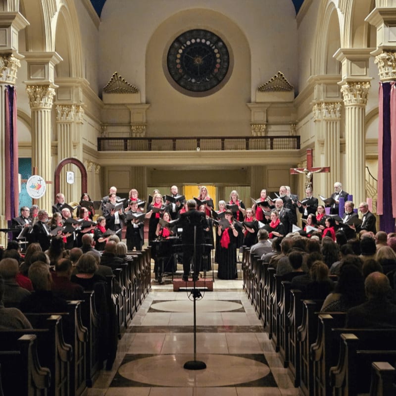 A choir performs in the foreground of a grand church, while an audience sits in pews before a backdrop of ornate columns and a large circular window.