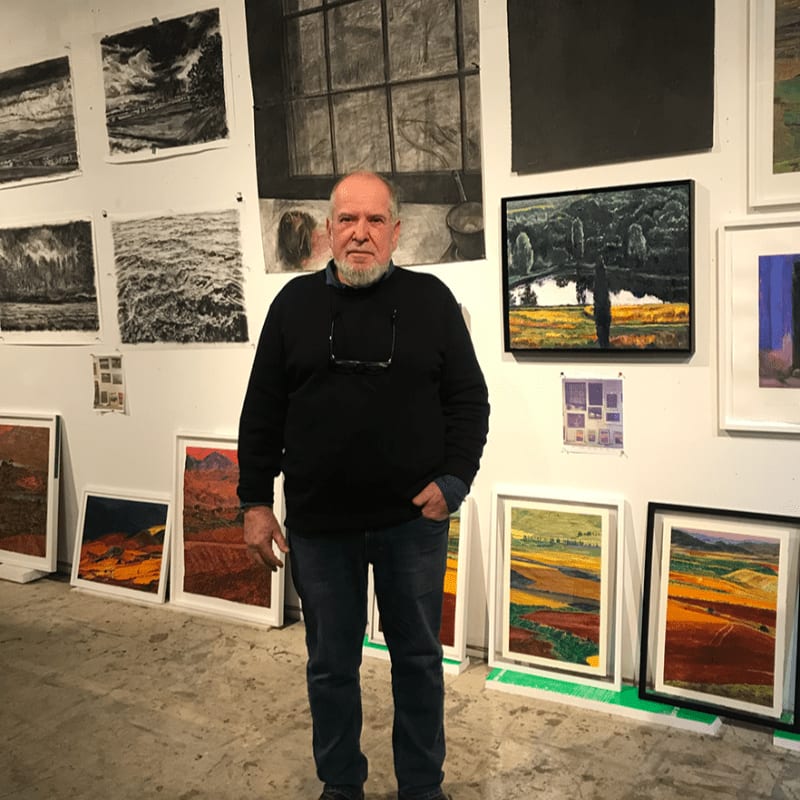 A bald man with a white beard stands in the foreground of an art gallery filled with various framed and unframed paintings on the walls and floor.
