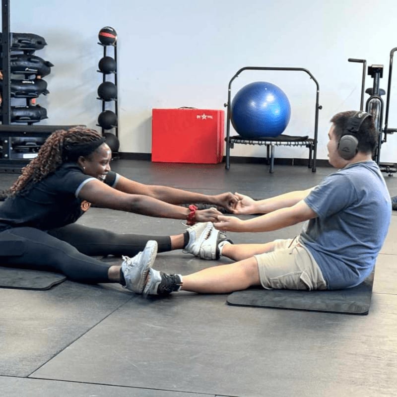 Two people sit facing each other and hold hands in a gym filled with exercise equipment and a blue stability ball.