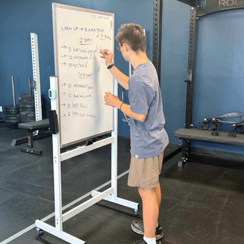 A young man writes a workout routine on a white board in a gym filled with weightlifting equipment.