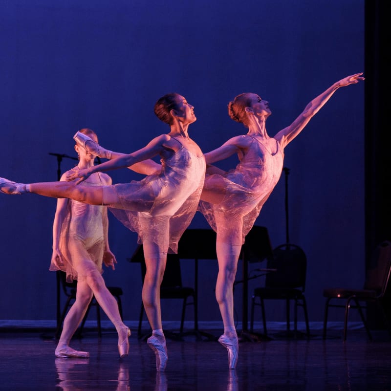 Three ballet dancers in sheer costumes perform on a dark stage with chairs and music stands in the background.