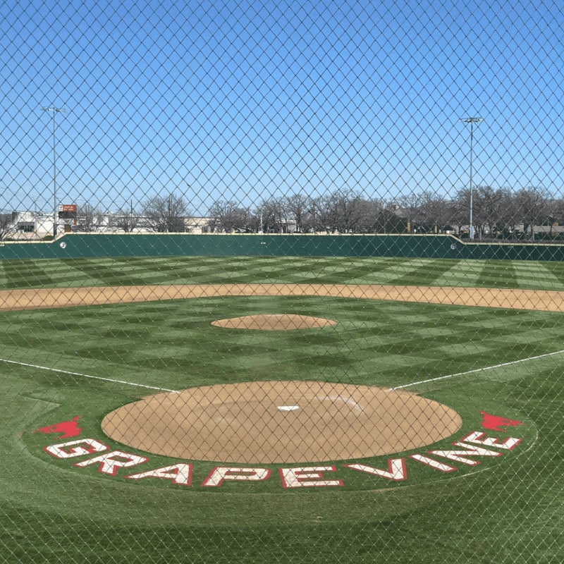 A baseball field with "GRAPEVINE" written in the dirt of the pitcher's mound is in the foreground, while a clear blue sky and distant trees form the background.