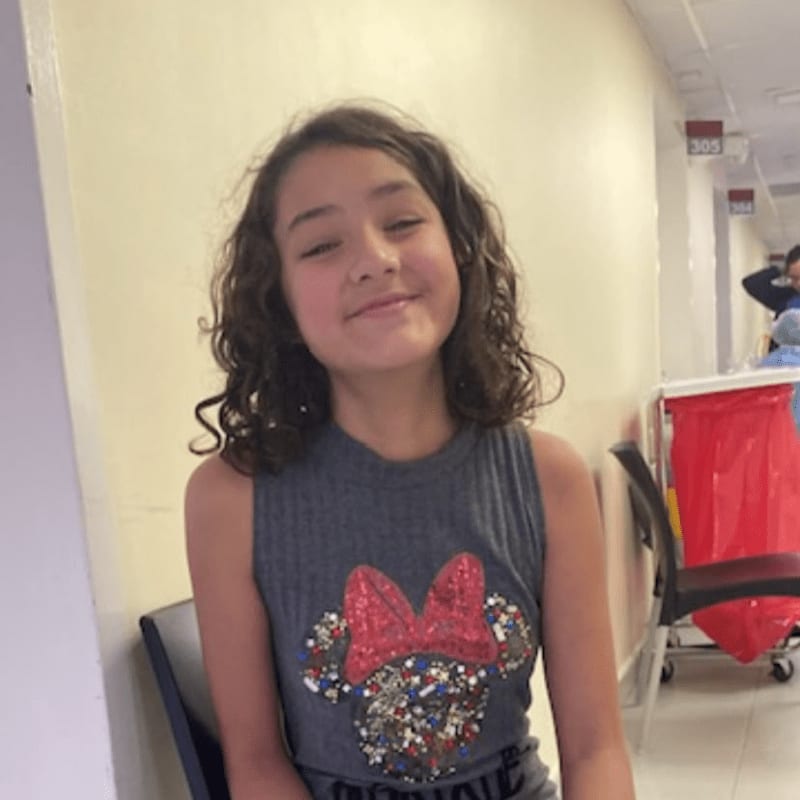 A young girl with curly hair wearing a Minnie Mouse shirt sits in a hallway with a red cart and room numbers in the background.