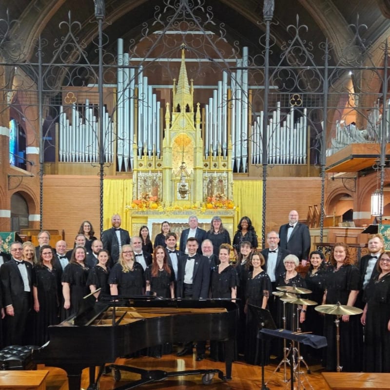 A choir stands in front of a grand pipe organ in a church, with a piano in the foreground.