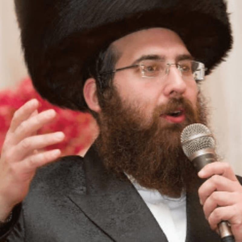A bearded man wearing a large black hat and glasses speaks into a microphone in the foreground against a blurred background with red flowers.