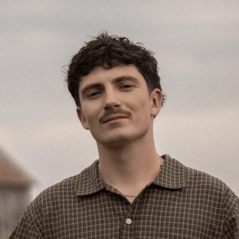A man with curly hair and a mustache wearing a checkered shirt stands against a plain, overcast sky.