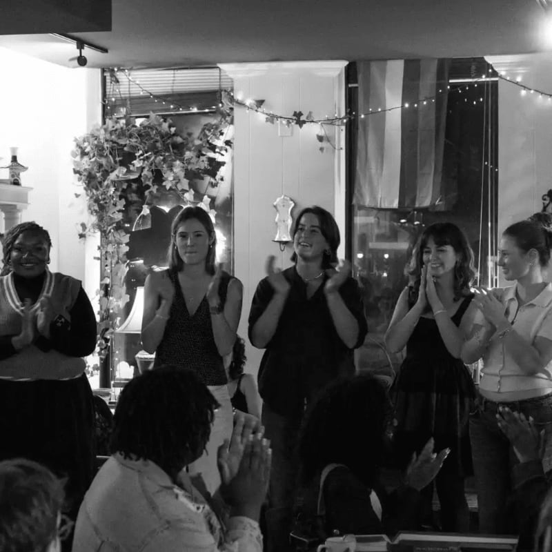 A group of people are clapping in the foreground of a room decorated with string lights and greenery in the background.