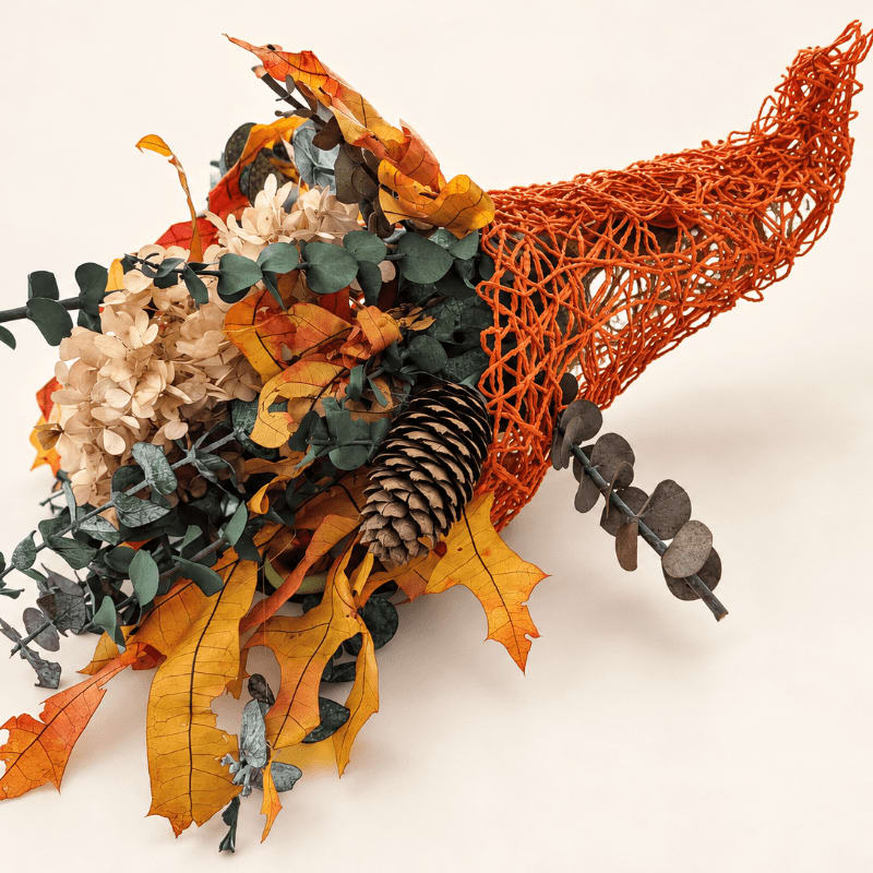 An orange woven cornucopia filled with autumn leaves, a pinecone, and dried flowers rests against a plain white background.