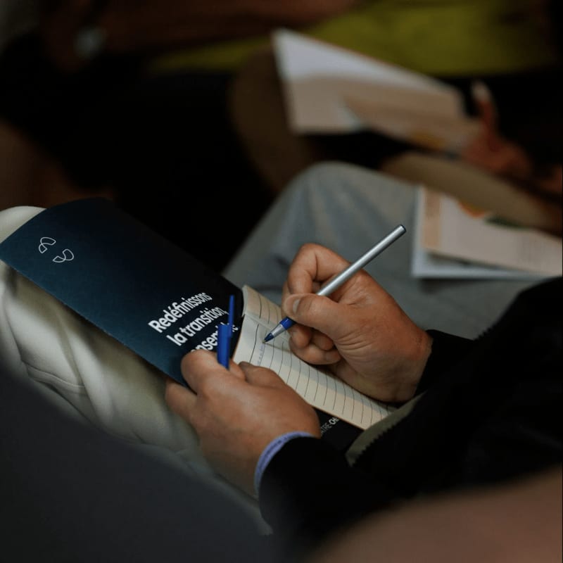 A person writes in a notebook with a silver pen in the foreground, while other people and papers are blurred in the background.