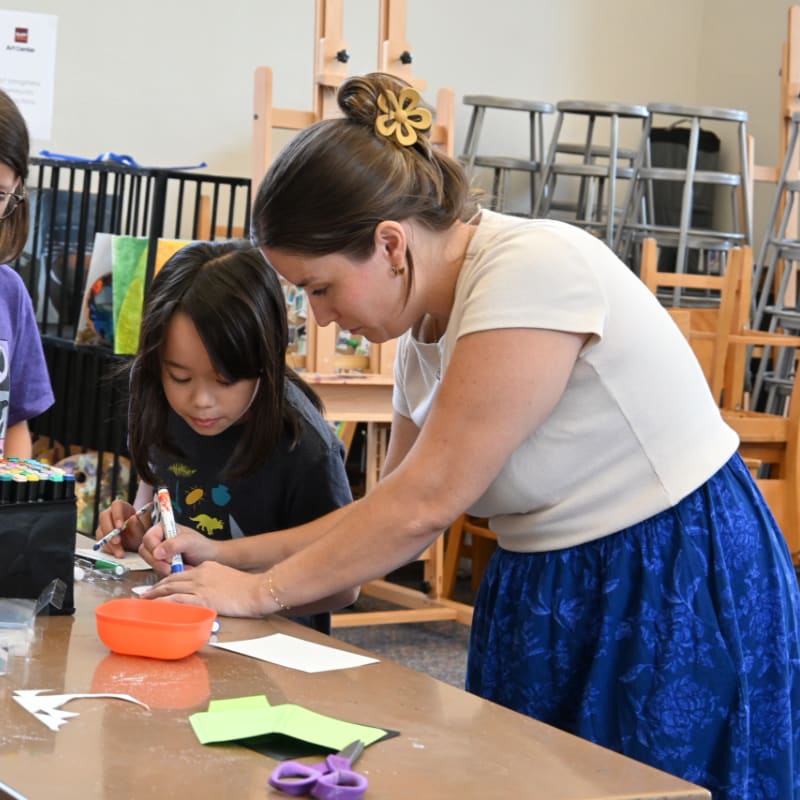 A woman in a blue skirt helps a young girl with an art project at a table in a classroom filled with chairs and easels.