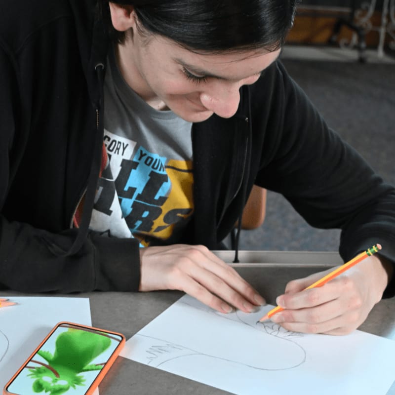 A person in a black hoodie draws on a piece of paper with a pencil in the foreground, while a blurred indoor setting forms the background.