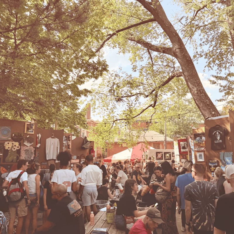 People browse outdoor art stalls under a canopy of large green trees.