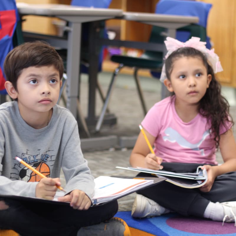 Two young children sit on a colorful rug holding pencils and notebooks in a classroom with desks and blue chairs in the background.