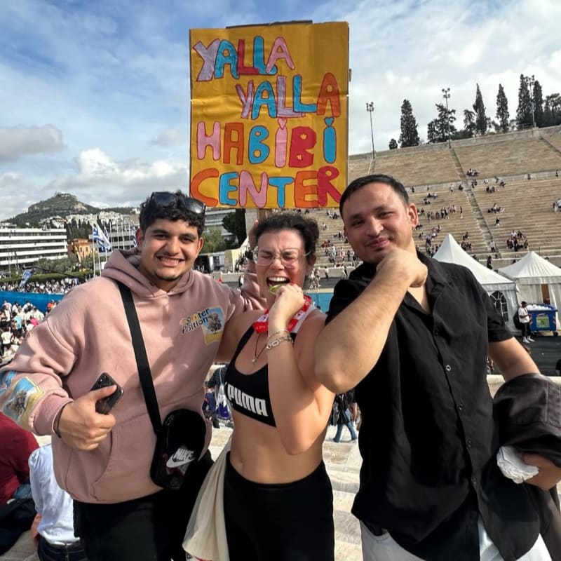 Three smiling people pose together in the foreground in front of a colorful "YALLA YALLA HABIBI CENTER" sign and a large ancient stone amphitheater.