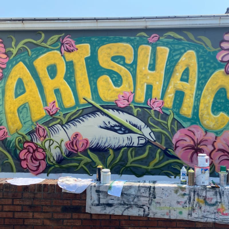 A colorful mural of a hand painting the words "ART SHACK" is painted on a wall above a brick ledge holding paint cans and supplies.