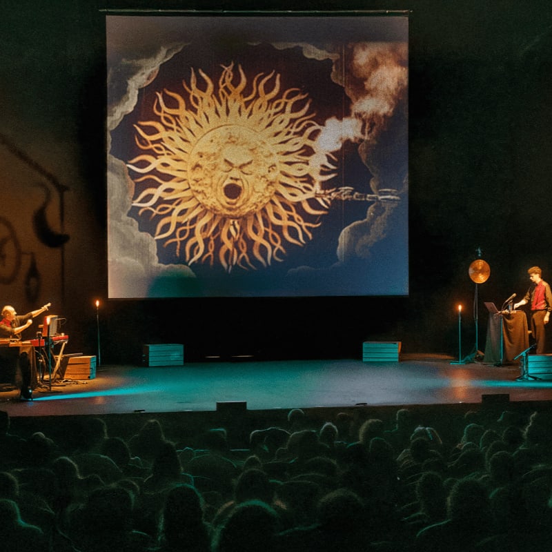 An audience sits in a dark theater facing a stage where performers stand before a large projection of a stylized, screaming sun.