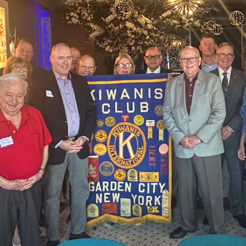 A group of men and women stand behind a blue Kiwanis Club banner in a room decorated with floral arrangements and sparkling lights.