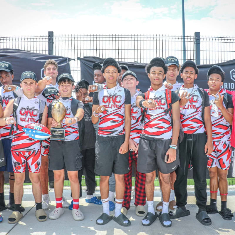 A group of young athletes in red and white striped uniforms pose with trophies in the foreground against a backdrop of black banners and a metal fence.