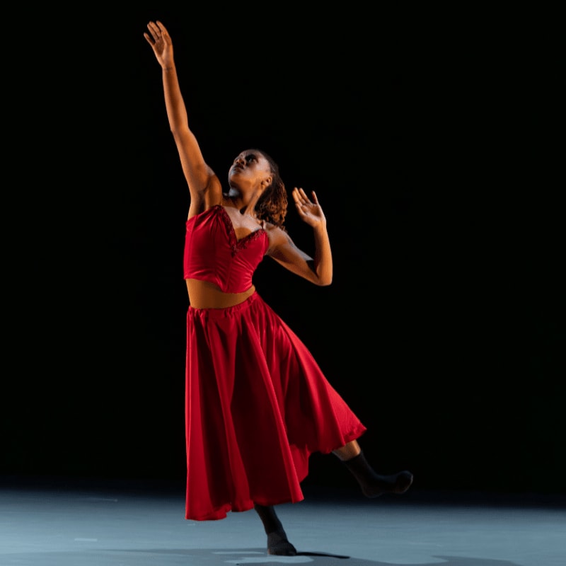 A woman in a red two-piece outfit dances with one arm raised against a stark black background.