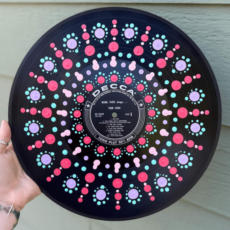 A hand holds a black vinyl record decorated with colorful pink, purple, and blue dots against a light-colored siding background.
