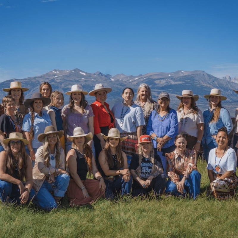 A group of women wearing cowboy hats and western attire pose together in a grassy field against a backdrop of distant mountains and a clear blue sky.