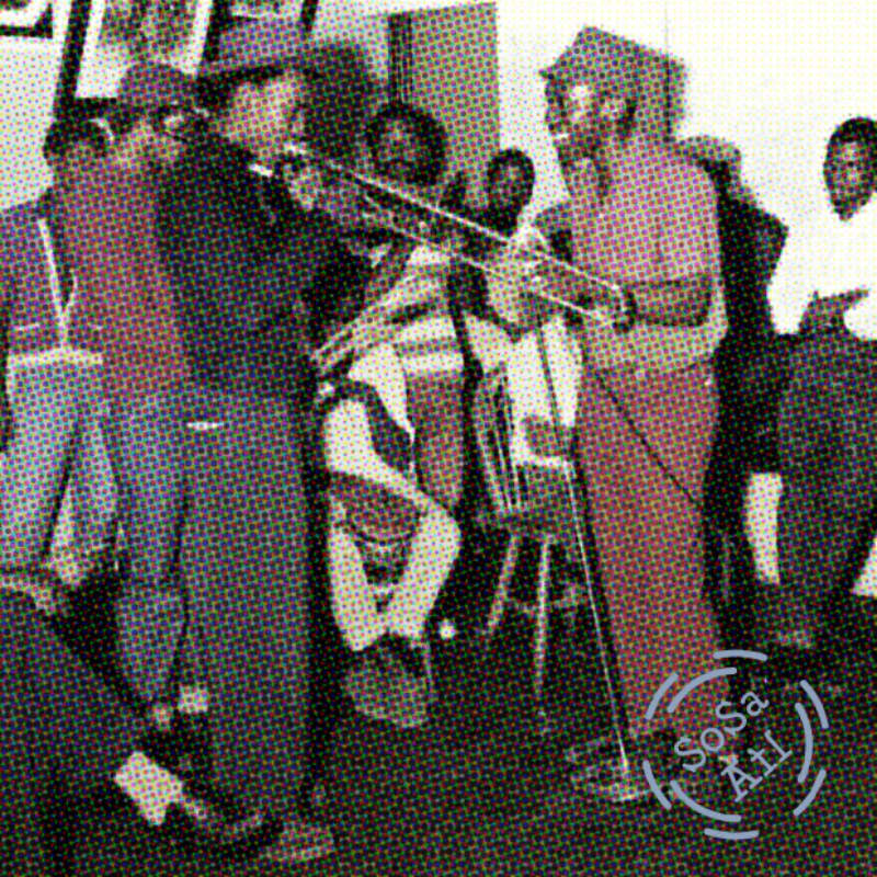 A group of men stand in a room, with two men in the foreground facing each other near a microphone stand.