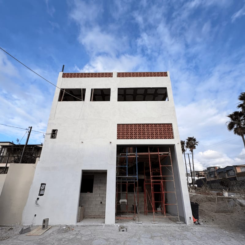 An unfinished white concrete building with scaffolding stands in the foreground against a bright blue sky with wispy clouds and distant palm trees.