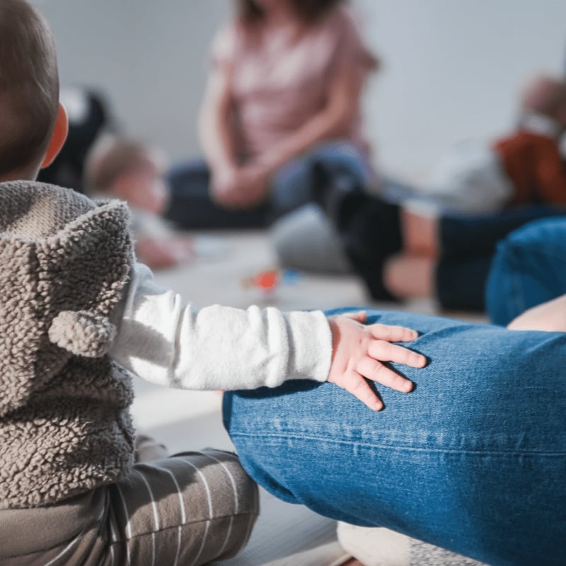 A small child in a fuzzy vest reaches out to touch a person's denim-clad leg, with other people blurred in the background.