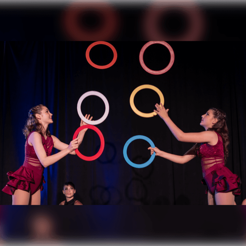 Two women in red dance costumes perform with colorful hoops against a dark curtained background.