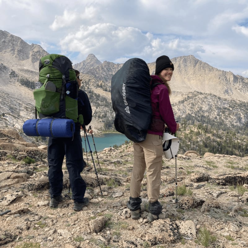 Two hikers with large backpacks stand on a rocky ridge overlooking a turquoise alpine lake and rugged mountains.