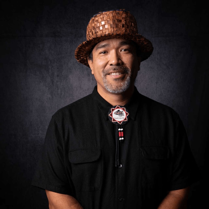 A smiling man wearing a woven brown hat and a black shirt with a decorative bolo tie stands against a dark, textured grey background.
