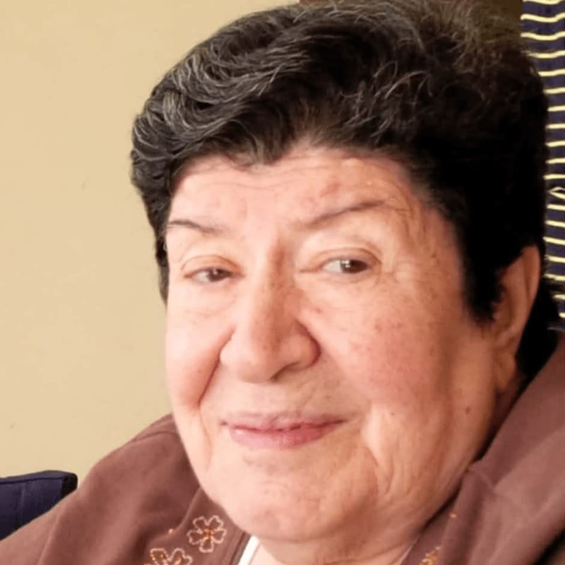 An elderly woman with short dark hair and a brown garment is positioned in the foreground against a plain, light-colored wall in the background.