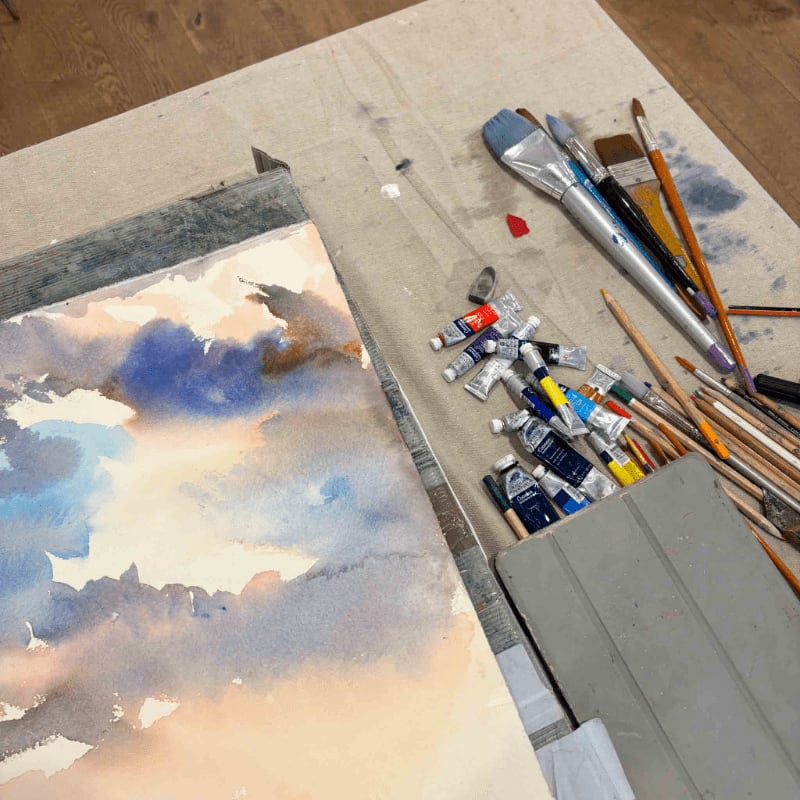 An abstract blue and white painting sits in the foreground next to a collection of paint tubes and brushes on a neutral-toned workspace.