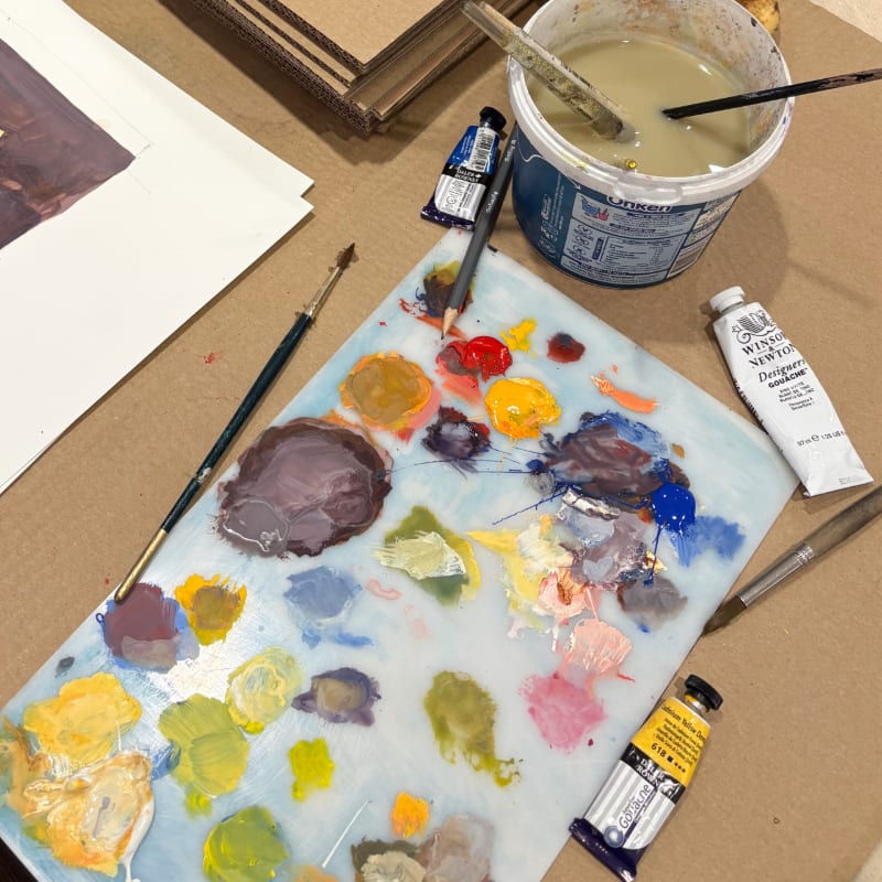 A paint-splattered palette and brushes sit in the foreground, with a bucket of water and art supplies in the background.