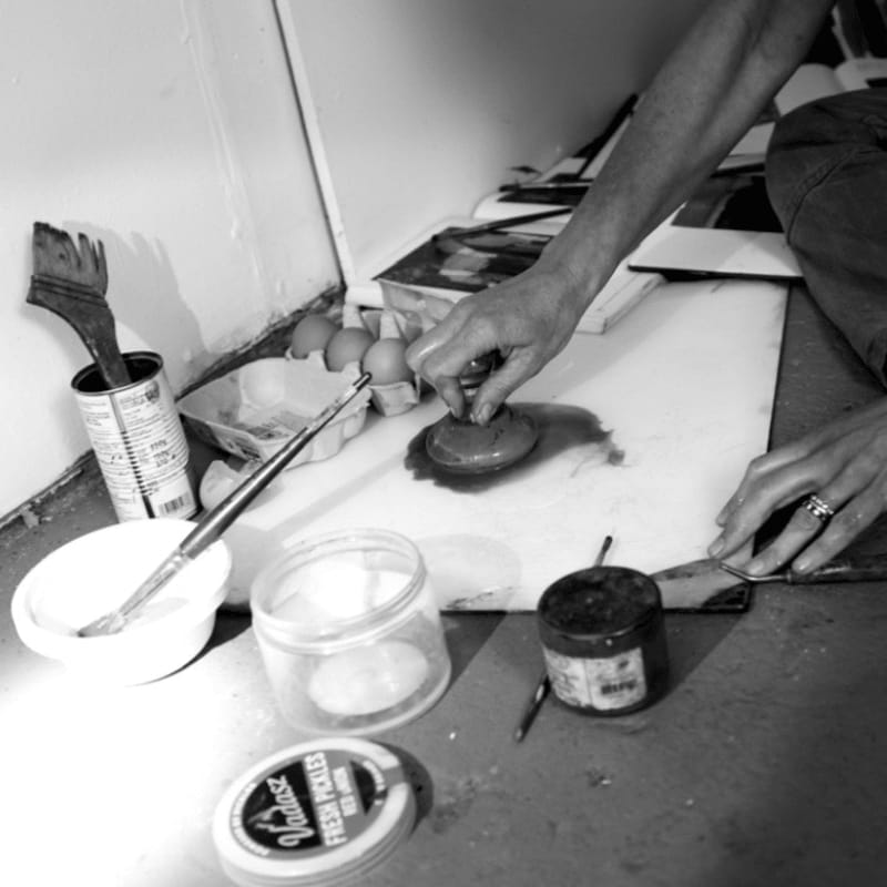 A person's hands work on a small clay object on a white surface, surrounded by art tools, eggs, and jars in a cluttered studio setting.