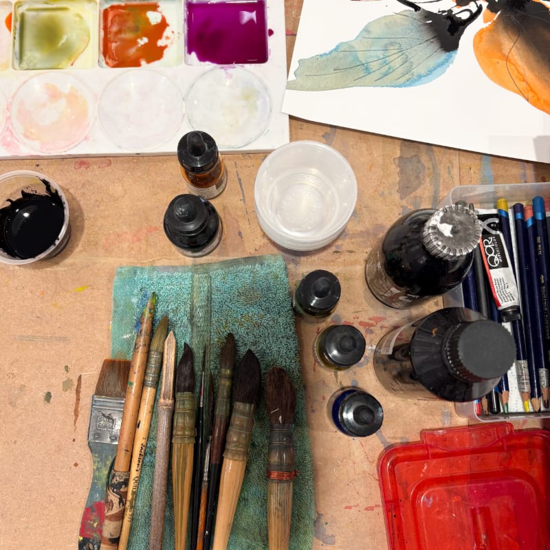 Various paintbrushes and ink bottles sit in the foreground on a paint-splattered table, with a watercolor palette and a painting in the background.
